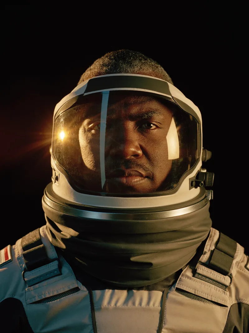 Dr. Yusuf Okonkwo in a flight helmet, coronal light across his visor